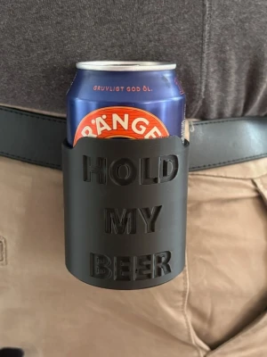 Svart bälthållare för burk - 2 för  80. Praktisk svart hållare som fästs i bältet och har texten 'HOLD MY BEER'. Perfekt för att ha en burk nära till hands på festival, grillkväll eller häng med vänner. Tillverkad i hård plast och passar de flesta standardburkar.