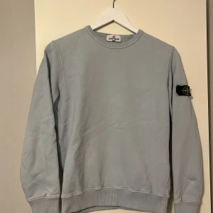 Ljusblå tröja från Stone Island - Säljer en stilren ljusblå tröja från Stone Island med deras ikoniska märke på ärmen. Tröjan har en rund hals och långa ärmar, perfekt för en avslappnad look. Passar bra till både jeans och chinos. Tröjan är äkta 