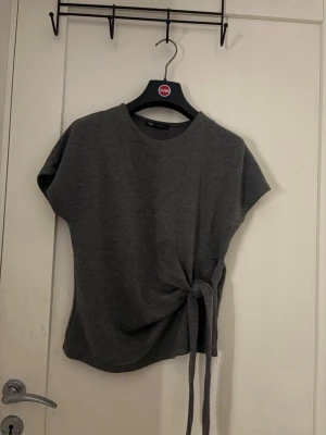 Grå topp med knyt i sidan - Snygg grå topp med korta ärmar och rund hals. Toppen har en unik detalj med knyt i sidan som ger en cool twist till din outfit. Perfekt att matcha med jeans eller kjol för en avslappnad men trendig look.