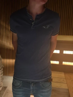 Mörkblå t-shirt med knappdetalj - Mörkblå t-shirt från Hollister med korta ärmar, knappdetalj vid halsen och vit logga på bröstet. Tillverkad i mjuk bomull och har kontrastsömmar vid ärmslut och nederkant för en clean look.