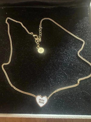 Guldigt hjärthalsband från Miu Miu - Snyggt halsband i guldfärgad metall från Miu Miu med ett hjärtformat hänge. Hänget har vit emalj på framsidan med Miu Miu-loggan och graverad baksida. Kedjan har en liten rund bricka med logga vid låset. Perfekt accessoar för att lyfta din outfit.