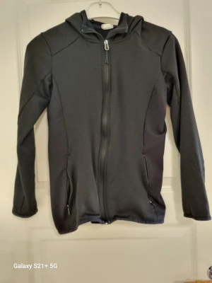Svart hoodie med dragkedja och huva - En stilren svart hoodie med hel dragkedja framtill och praktisk huva. Tröjan har två fickor med dragkedja och är tillverkad i ett mjukt material som känns skönt mot huden. Perfekt för dig som gillar en enkel och clean look.