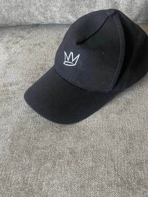 Svart keps med vit krona brodyr - Snygg svart keps med böjd skärm och en stilren vit broderad krona framtill. Klassisk modell med sex paneler och knapp på toppen. Tillverkad i ett slitstarkt tyg som känns bekvämt och passar till de flesta outfits.