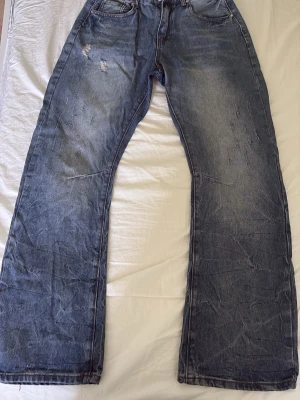 Blåa baggy jeans med slitningar - Säljer ett par blåa bootcut jeans från peaceoflifestudio, vida ben och slitna detaljer på framsidan. Jeansen har klassiska fem fickor och en tvättad look som ger en avslappnad vibe. Perfekta för dig som gillar streetwear och oversized stil.