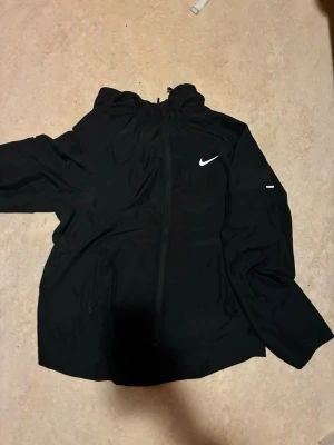 Nike Miller - Snygg svart vindjacka från Nike med dragkedja framtill och huva. Jackan har en vit Nike-logga på bröstet och diskreta fickor på sidorna. Perfekt för blåsiga dagar och enkel att matcha med olika outfits.