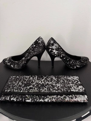 Svarta pumps med paljetter och matcha de väska - Snygga svarta pumps täckta med glittrande silverpaljetter och smal klack. Perfekta för att sticka ut på festen eller när du vill ha extra bling. Rundad tå och klassisk siluett. Matchande clutch i samma material och stil ingår.