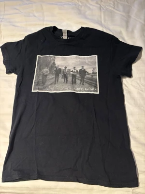 Svart t-shirt med tryck  - Svart t-shirt med ett stort svartvitt tryck på bröstet som visar fem personer i kostym och texten 'L'ARTE Est. 2023'. Klassisk passform och rund hals. Perfekt för dig som gillar stilrena motiv och streetwear-vibe. Bara att höra av sig vid funderingar!