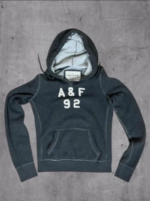 Mörkgrå hoodie Abercrombie & Fitch - Mörkgrå hoodie med huva och magficka från Abercrombie & Fitch. Framtill står det 'A&F 92' i vita bokstäver. Hoodien har ribbade muddar och kontrastsömmar samt mjukt innerfoder. Perfekt för en avslappnad och sportig stil. Kan diskutera pris🤝