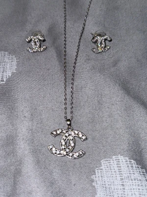 Set med halsband och örhängen i silver - Säljer ett matchande set med halsband och örhängen i silverfärg med glittrande stenar. Båda smyckena har en ikonisk logga som hängsmycke och studs. Perfekt för dig som gillar stilrena accessoarer med extra bling.