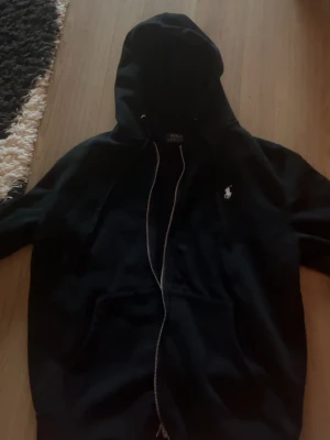 Svart hoodie från Polo Ralph Lauren - Svart hoodie med dragkedja från Polo Ralph Lauren. Klassisk design med huva och dragsnören, samt vit broderad logga på bröstet. Mjuk bomullskänsla och känguruficka framtill. Perfekt för en chill och stilren look.