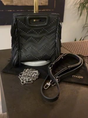 Söt mini Maje väska - Snygg svart liten väska från Maje med dekorativa silver mönster. Detaljerna är något rostiga längst ner med inget som märks!! Tillkommer även dustbag, kedja och ett justerbart axelremband som tillhör väskan❤️😊
