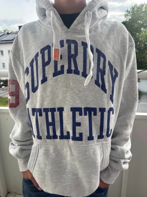 Gråvit  Superdry hoodie med tryck - Snygg grå hoodie från Superdry med stor blå text 'SUPERDRY ATHLETIC' på framsidan och röd '49' på ärmen. Klassisk känguruficka, vita dragsnören och huva. Storlek XL men superdry har ganska små storlekar
