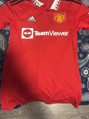 Manchester United matchtröja Adidas XS - Röd Manchester United matchtröja från Adidas med klubbmärke, TeamViewer-logga och klassisk vit krage med röda detaljer. Tröjan har svarta axelränder och är tillverkad i lätt funktionsmaterial, perfekt för fotbollsträning eller support.