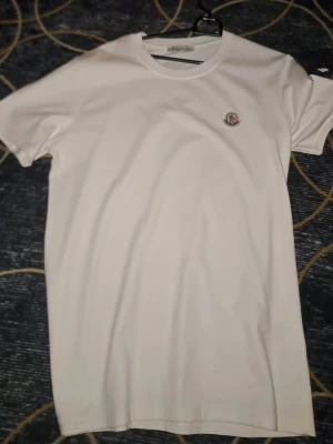 Vit t-shirt från Moncler - Säljer en stilren vit t-shirt från Moncler med klassisk rund hals och Moncler-logga broderad på bröstet. T-shirten är kortärmad och gjord i mjuk bomull för maximal komfort. Perfekt till jeans eller shorts för en clean look.