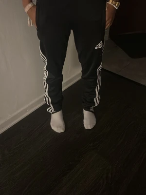 Svarta Adidas mjukisbyxor med vita ränder - Svarta mjukisbyxor från Adidas med klassiska vita ränder längs sidorna och Adidas-logga på låret. Byxorna har en avslappnad passform och resår i midjan. Perfekta för en sportig och chill stil.