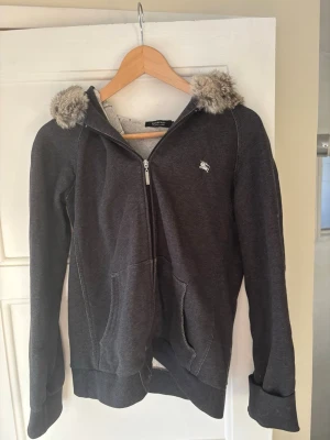Exklusiv vintage Burberry Black Label hoodie/jacka  - Svårhittad modell från Burberry Black Label, en Japan-specifik Burberry-linje som lanserades 1998 och fanns fram till 2015. Eftersom linjen inte längre produceras är äldre modeller idag extra eftertraktade bland vintage- och designerälskare.  Stilren mörkblå/grå design med Burberry-emblem på bröstet, dragkedja, två fickor och exklusiva pälsdetaljer på huvan. Modellen är unisex och passar både dam och herr.