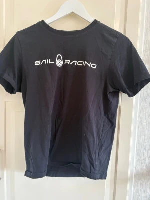 Svart Sail Racing t-shirt - Svart t-shirt från Sail Racing med vit logga och text på ryggen. Klassisk rund hals och korta ärmar. Tillverkad i mjuk bomull som känns skön mot huden. Perfekt för en avslappnad och sportig stil.