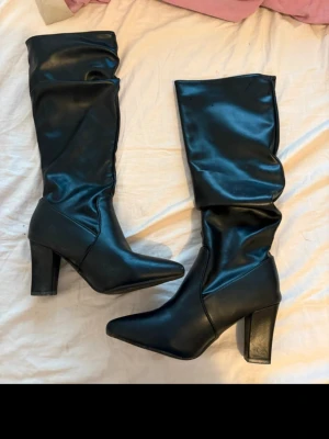 Svarta höga stövlar med klack - Säljer ett par svarta höga stövlar i glansigt konstläder med bred klack och spetsig tå. Stövlarna har en slouchy passform upptill och ger en cool och trendig look. Perfekta till både jeans och kjol för en edgy stil.