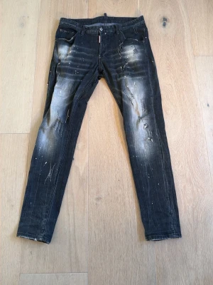 Svarta dsquared jeans - Säljer ett par svarta dsquared jeans med coola slitningar och blekta detaljer. Jeansen har en smal passform, klassisk femficksmodell och vita färgstänk för en trendig look. Perfekta för dig som gillar streetstyle och vill sticka ut.