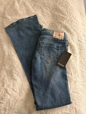 Blå bootcut jeans från True Religion - Snygga blå bootcut och lågmidjade jeans från True Religion Jeansen har en ljus tvätt och har rosa detaljer både fram och baktill. Jeansen är aldrig använda pga att det är fel storlek💕 Prislappen är kvar och jeansen är i storlek w27, jag säljer då dom e för stora