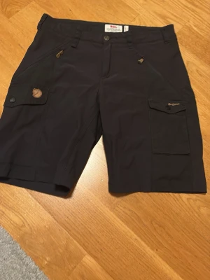 Svarta shorts från Fjällräven dam - Snygga svarta shorts från Fjällräven med flera praktiska fickor och dragkedjor. Modellen har normal passform och är tillverkad i slitstarkt material, perfekta för utomhusaktiviteter. Klassisk design med knappstängning och detaljerade logotyper.
