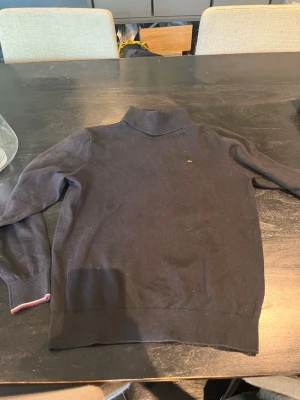 Svart polotröja Tommy Hilfiger - Säljer en svart stickad polotröja från Tommy Hilfiger. Tröjan har en liten broderad logga på bröstet och ribbade muddar vid ärmslut och nederkant. Klassisk och stilren design, perfekt till höst och vinter.