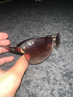 Ray-Ban pilotbågar i guld och brunt - Snygga Ray-Ban solglasögon med klassiska pilotbågar i guld och bruna tonade glas. Bågarna är i metall och har Ray-Ban-loggan på linsen och sidan. Perfekt accessoar för soliga dagar och ger en cool vibe till din outfit.