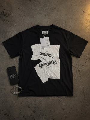 Maison Margiela svart t-shirt - Säljer en svart t-shirt från Maison Margiela med ett stort vitt grafiskt tryck och texten 'maison margiela' på framsidan. Klassisk rund hals och korta ärmar. Tillverkad i mjuk bomull för en skön känsla.