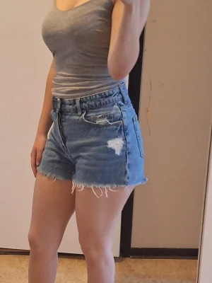 Blå jeansshorts - Säljer ett par blå jeansshorts med råa, fransiga kanter och slitningar på framsidan. Klassisk femficksmodell i denim med knappar istället för dragkedja. Perfekta för sommaren och ger en avslappnad vibe.