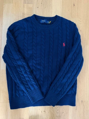 Mörkblå kabelstickad tröja Polo Ralph Lauren - Mörkblå kabelstickad tröja från Polo Ralph Lauren med rund halsringning och röd broderad logga på bröstet. Tröjan har ribbade muddar vid ärmslut och nederkant, perfekt för en klassisk och stilren look.