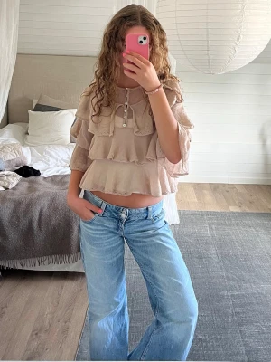 Beige volangblus med knappar - Supersöt beige blus med flera volanglager och knappar framtill. Croppad modell med trekvartsärm och rund halsringning. Supersnygg att styla med ett par basic jeans eller en fin kjol! Från Zara i storlek M.