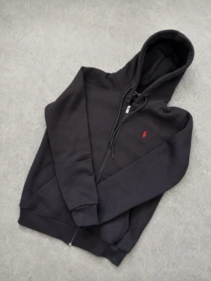 Ralph Lauren Ziptröja  - Säljer en svart Ralph Lauren hoodie med dragkedja och huva. Klassisk design med fickor framtill och en liten röd logga broderad på bröstet. Hoodien har ribbade muddar vid ärmslut och nederkant samt justerbara snören i huvan. Perfekt för en chill och stilren look. Har från storlek L-XXL