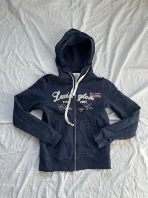 Marinblå Lexington zip-hoodie - Snygg marinblå hoodie från Lexington med dragkedja, vit broderad logga och amerikansk flagga på bröstet. Mjuk bomullskvalitet, vita snören i huvan och kängurufickor framtill. Perfekt för en avslappnad och stilren look.