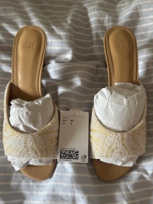 Beige pärlbroderade pumps från H&M - Säljer ett par snygga pumps från H&M i storlek 37. Skorna är beige med öppen tå och dekorerade med vita och ljusgula pärlor i ett mönster. De har en låg klack och öppen häl, vilket ger en trendig look. Perfekta för att lyfta din outfit!