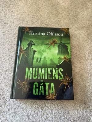 Mumiens gåta av Kristina Ohlsson - En spännande bok med hård pärm och grönt omslag, skriven av Kristina Ohlsson. Omslaget har silhuetter av gravstenar, spindlar och mystiska figurer, vilket ger en kuslig känsla. Perfekt för dig som gillar mysterier och äventyr inom skönlitteratur.