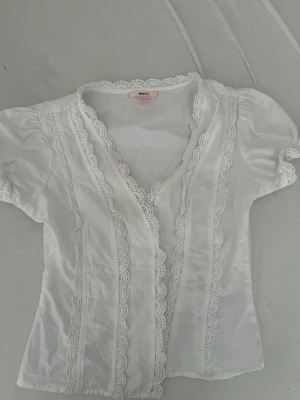 Vit blus med spetsdetaljer - Söt vit blus med korta puffärmar och dekorativa spetskanter längs v-ringningen och framtill. Blusen har en feminin passform och romantiska detaljer som ger en vintagekänsla. Perfekt att styla med jeans eller kjol för en trendig look.