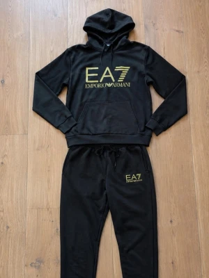 Svart EA7 Emporio Armani Tracksuit - Snygg svart tracksuit från EA7 Emporio Armani med guldfärgad logga på både tröja och byxor. Setet består av en hoodie med känguruficka och matchande byxor med dragsko och muddar vid benslut. Perfekt för chill dagar hemma eller när du vill vara bekväm. Storlek S