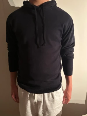 Mörkblå Ryvls hoodie - En stilren mörkblå hoodie med huva och dragsko Hoodien har långa ärmar, ribbade muddar vid ärmslut och nederkant samt en mjuk, slät yta. Perfekt för en avslappnad look och enkel att matcha med olika outfits.