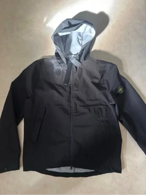 Svart vindjacka från Stone Island - Snygg svart vindjacka från Stone Island med ikonisk patch på ärmen. Jackan har huva, dragkedja framtill och två snedställda fickor. Materialet är tunt och vindtätt, perfekt för blåsiga dagar. Stilsäker och enkel att matcha till olika outfits.