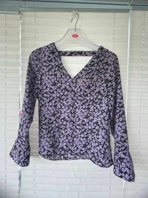 Lila blommig blus från Gina Tricot - Snygg blus från Gina Tricot i svart med lila blommönster. Blusen har v-ringning och långa ärmar med lätt utsvängda ärmslut. Tunt och luftigt material som ger en skön känsla. Perfekt för dig som gillar trendiga mönster och en avslappnad look.