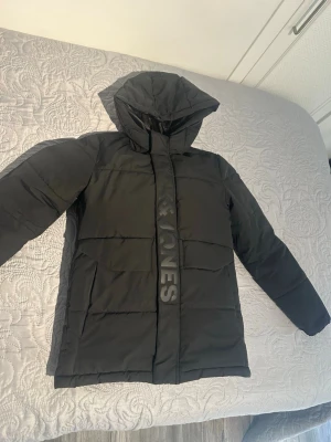 Svart pufferjacka med huva - Snygg svart pufferjacka med stor huva och diskret logga på framsidan. Jackan har raka ärmar, quiltad design och dragkedja med vindslå. Perfekt för kalla vinterdagar och ger en modern look. Tillverkad i slitstarkt syntetmaterial.