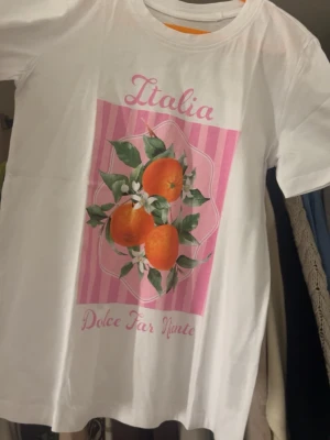 Vit t-shirt med apelsinmotiv och text - Säljer en vit t-shirt i storlek XS med färgglatt apelsinmotiv och texten 'Italia' och 'Dolce Far Niente' i rosa. T-shirten har korta ärmar och är gjord i mjuk bomull. Perfekt för dig som gillar fräscha prints och vill ha en chill vibe. Passar en S/M då den är stor i storleken. Köptes i Grekland så vet tyvärr ej märke.