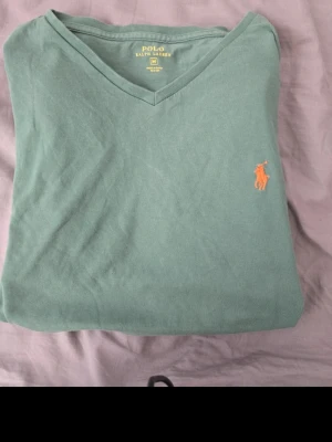 Grön v-ringad t-shirt från Polo Ralph Lauren - Snygg grön t-shirt från Polo Ralph Lauren med v-ringning och den klassiska orange broderade loggan på bröstet. Tillverkad i mjuk bomull och har en clean, stilren look som passar perfekt till jeans eller shorts.