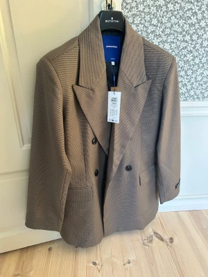 Brun oversized kavaj från Jack & Jones - Snygg brun oversized kavaj från Jack & Jones med dubbelknäppning och breda slag. Kavajen har ett diskret rutmönster och två framfickor. Perfekt för en trendig och avslappnad look. Materialet är mjukt och följsamt. Helt oanvänd. Storlek S men stor så passar bättre M. Nypris 1299kr. 