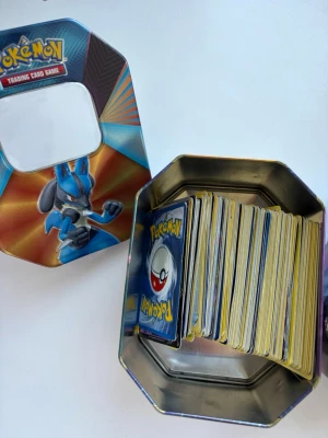 Pokémon samlarkort i plåtlåda - Säljer en samling Pokémonkort i en färgglad plåtlåda med motiv av Lucario och Pikachu. Lådan är åttakantig och har ett fönster på locket. Perfekt för dig som gillar Pokémon och vill utöka din samling med många olika kort.