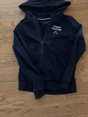 Svart hoodie från Tommy Hilfiger - Snygg svart hoodie från Tommy Hilfiger med dragkedja framtill och huva. Tryck med logga och 'Est. 1985' både fram och bak. Klassisk design med ribbade muddar och fickor framtill. Perfekt för en avslappnad och stilren look. MAN KAN ÄVEN FÅ MED BYXOR 550kr för hela setet!!