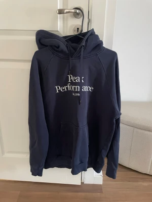 Mörkblå Peak Performance hoodie - Snygg mörkblå hoodie från Peak Performance med vit logga och text på bröstet. Klassisk känguruficka framtill, justerbar huva med dragsko och mjukt material. 