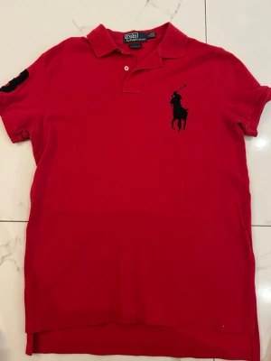 Röd pikétröja från Ralph Lauren - Klassisk röd pikétröja från Ralph Lauren med stor svart broderad logga på bröstet och krage med knappslå. Tröjan har korta ärmar och gjord i pikématerial.