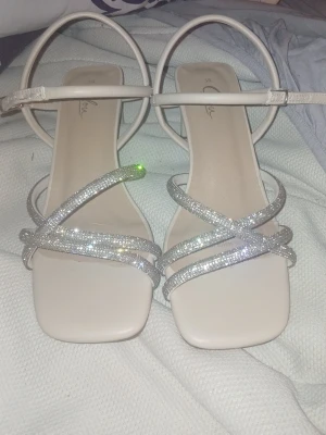Glittriga vita sandaler från Clou - Säljer ett par vita sandaler från Clou i storlek 38. De har fyrkantig tå, tunna remmar med silverglitter och justerbart spänne runt ankeln. Perfekta för dig som vill ha något extra till outfiten. Materialet är syntet med glittriga detaljer.