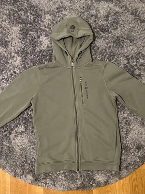 Grön hoodie från Sail Racing - Snygg grön hoodie från Sail Racing med dragkedja framtill och en extra ficka med dragkedja på bröstet. Tröjan har huva med justerbara snören och ribbade muddar. Perfekt för dig som gillar en clean och sportig stil. Den har inga deffekter så hör av dig om du har några frågor.